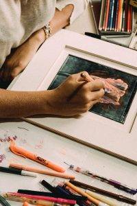 Photo de près d'un bureau d'artiste, une main qui tient un crayon, la jeune femme est en train de dessiner une magnifique fleur dans les tons verts et roses.